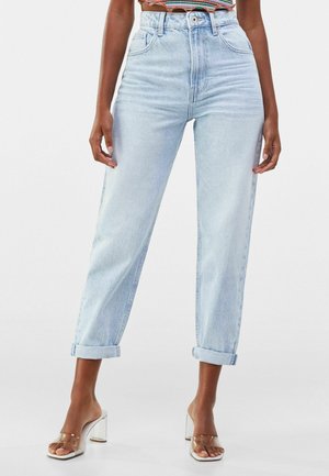 Woman wearing light blue high-waist rolled-up jeans and clear heeled sandals, standing with hands relaxed by sides.