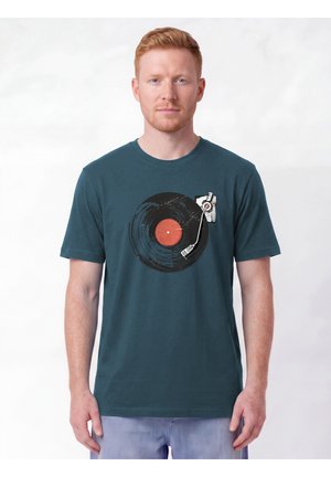 Homme aux cheveux roux courts portant un t-shirt bleu sarcelle avec une image d'un disque vinyle noir et d'une platine sur fond blanc.