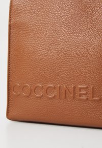 Tan leather wallet with textured surface, featuring embossed "COCCINEL" logo, smooth edges, and a minimalist design.