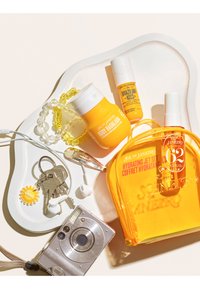 Produits de soin pour la peau Sol de Janeiro jaunes, une pochette orange transparente, des clés avec un porte-clés soleil, des écouteurs blancs et un appareil photo argenté sur une surface claire.