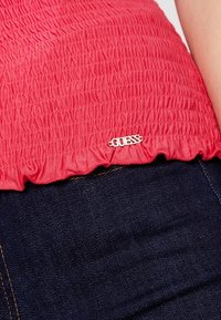 Rød smocket top med en bølget kant, der har et sølvfarvet "GUESS" logo mærke. Kombineret med mørk denimstof i nærheden.