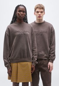 Sudaderas marrones con detalle bordado "TOMMY JEANS", combinadas con una falda amarilla de cuadros. Corte casual con puños y bajo acanalados.