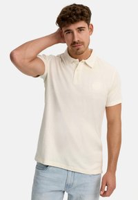 Helles cremefarbenes Polo-Shirt mit kurzen Ärmeln, Drei-Knopf-Blende und dezentem Logo-Detail auf der Brust, kombiniert mit hellblauen Jeans.