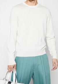 Biały sweter z długim rękawem, z okrągłym dekoltem i subtelnym teksturowanym wzorem, zestawiony z zielonkawymi spodniami i biało-czarną torebką.