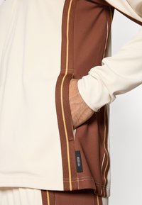 Creme lange mouw top met bruine zijkanten en dunne gele strepen. Stof lijkt textuur te hebben met zichtbare stiksel en een merklabel.