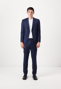 Traje navy con corte ajustado, chaqueta de un solo pecho, dos botones y pantalones a juego. Combinado con una camiseta blanca y zapatos negros.