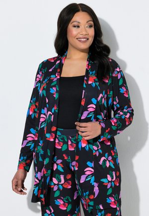 Kvinde smilende, iført sort top med matchende cardigan med blomsterprint og bukser i røde, lyserøde, blå, grønne og lilla farver.
