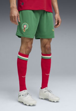 Beine mit grünen Shorts mit Marokko-Emblem, roten Socken mit grünen und weißen Streifen und weißen Puma Fußballschuhen auf grauem Hintergrund.