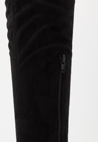 Czarny zamszowy but za kolano z teksturowaną powierzchnią i pionowym czarnym zamkiem błyskawicznym po zewnętrznej stronie.