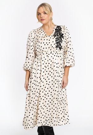 Femme portant une robe midi en satin crème avec des pois noirs et une appliqué en dentelle florale noire sur l'épaule, associée à des bottes noires.