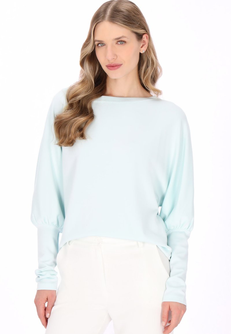 Jonge vrouw met lang golvend haar, gekleed in een lichtblauw shirt met lange mouwen en witte broek, staand tegen een effen witte achtergrond.