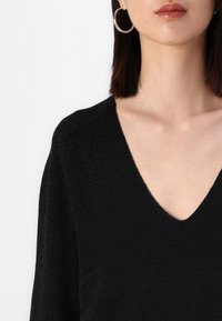 Femme portant un pull noir côtelé à col en V et de grandes boucles d'oreilles argentées, avec des cheveux longs et lisses et un gloss naturel sur les lèvres.