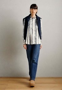 Button-up skjorte med vertikale marineblå og hvite striper, kombinert med en marineblå vest og blå jeans. Skoene er beige klumper. Bakgrunn av tregulv.