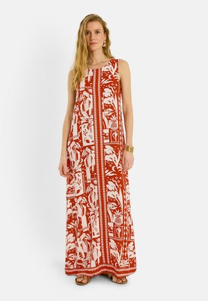 Mujer con vestido sin mangas, largo hasta el suelo, con estampado rojo y blanco, con pulsera y pendientes de oro, de pie frente a un fondo blanco.