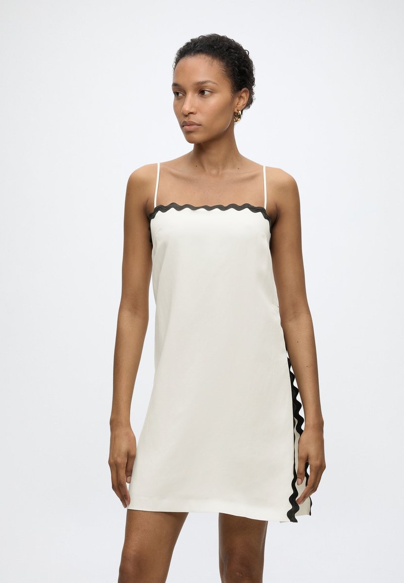 Γυναίκα που φοράει λευκό αμάνικο φόρεμα με λεπτούς ιμάντες και μαύρη κυματιστή διακόσμηση κατά μήκος της λαιμόκοψης και της πλευράς, στέκεται μπροστά σε λευκό φόντο.