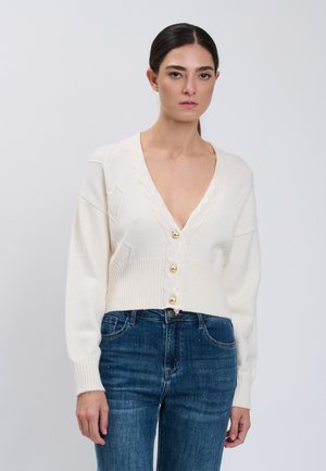 Frau trägt einen weißen V-Ausschnitt-Cardigan mit goldenen Knöpfen und blauen Jeans und steht vor einem einfarbigen hellgrauen Hintergrund.