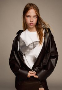 Hnědá kožená oversized bunda přes bílý tričko s černým motivem motýla. Model má ruce sepnuté v pase.