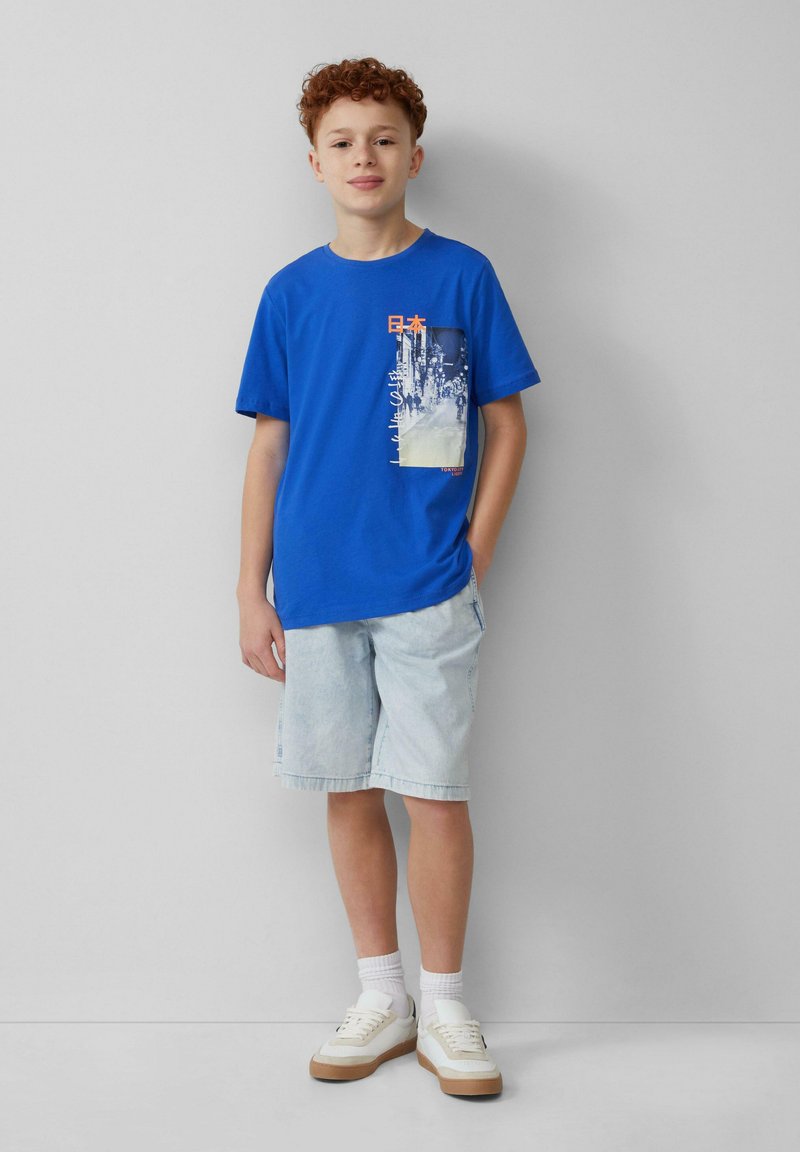 Blauwe katoenen t-shirt met een grafisch zakontwerp met Japanse tekst, gecombineerd met lichte denim shorts en witte sneakers.