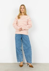 Femeie purtând un pulover tricotat cu dungi roz, blugi largi albaștri și pantofi cu toc maro, cu vârful ascuțit, stând în fața unui fundal alb.