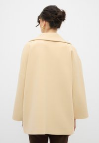 Cappotto oversize beige con una texture liscia, maniche ampie e colletto classico. Il retro presenta dettagli delle cuciture sottili e una finitura pulita.