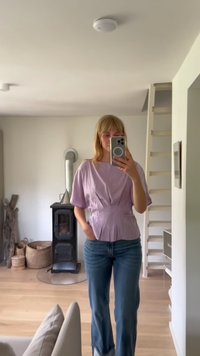 Helles lila, kurzärmlige Bluse mit plissiertem Vorderteil, kombiniert mit blauen Denim-Jeans. Glatte Textur, figurbetontes Design und lässige Silhouette.