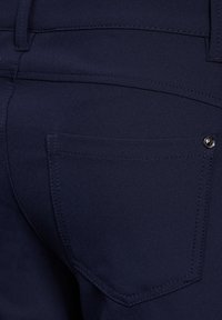 Tmavě modré kalhoty s kapsou na zadní straně, zdobenou švy a stříbrným knoflíkem. Materiál vypadá hladce a mírně strukturovaně.