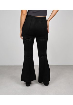 Mujer con pantalones negros acampanados de talle alto y una camiseta corta oscura, de pie con una mano en la cadera contra un fondo liso.