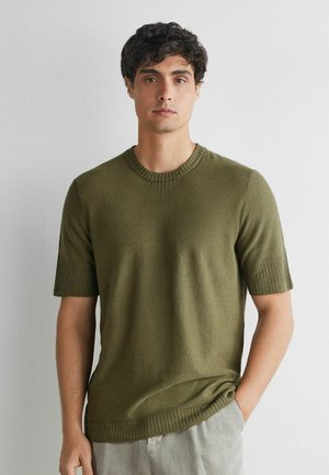Young man with dark curly hair wears olive green short-sleeve sweater and light gray pants, standing against a plain white wall.