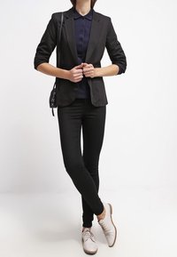 Veste noire sur un polo bleu marine, associée à un jean skinny noir et des chaussures lacées gris clair. Design simple avec une coupe ajustée et un look sur mesure.