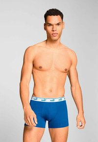 Shirtless man with short curly hair wearing blue Puma boxer briefs with white waistband, standing against a plain light background.