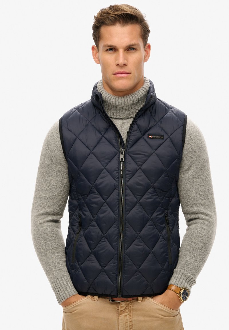 Marineblauwe gewatteerde gilet met een hoge kraag, een rits aan de voorkant en zijzakken; gedragen over een grijze geribde coltrui.