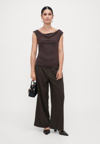 Woman standing against plain background wearing a sleeveless brown top, pinstriped wide-leg pants, black heels, and holding a small black handbag.