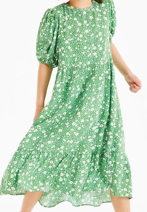 Robe florale verte en tissu léger, présentant des manches ballons, un ourlet en couches et un motif de fleurs blanches dispersées.