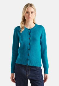 Giovane donna con capelli biondi lisci che indossa un cardigan teal abbottonato e jeans blu scuro su uno sfondo neutro.