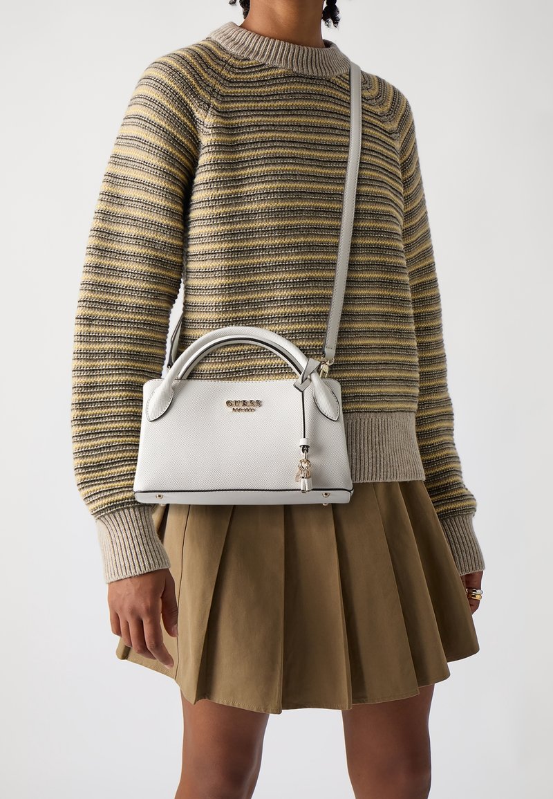 Personne portant un pull rayé et une jupe plissée marron, portant un petit sac à main blanc Guess avec des poignées supérieures et une bandoulière.
