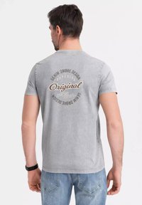 Šedé tričko s krátkým rukávem s potiskem v retro stylu na zádech, obsahující kruhový text v béžové a hnědé barvě.