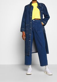 Denim lång rock med kontrasterande sömmar, två framfickor, i kombination med en gul kort topp och högmidjade blå jeans. Vita sneakers.