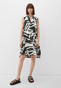 Schwarz-weißes ärmelloses Kleid mit Kragen, das ein Blattmuster, einen V-Ausschnitt und eine ausgestellte Silhouette aufweist. Getragen mit schwarzen Sandalen.