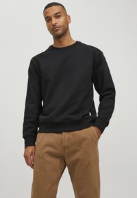 Schwarzer Sweatshirt mit Rundhalsausschnitt, gerippten Bündchen und Saum. Kombiniert mit hellbraunen Hosen. Glatte Textur, lässige Passform, minimales Branding.