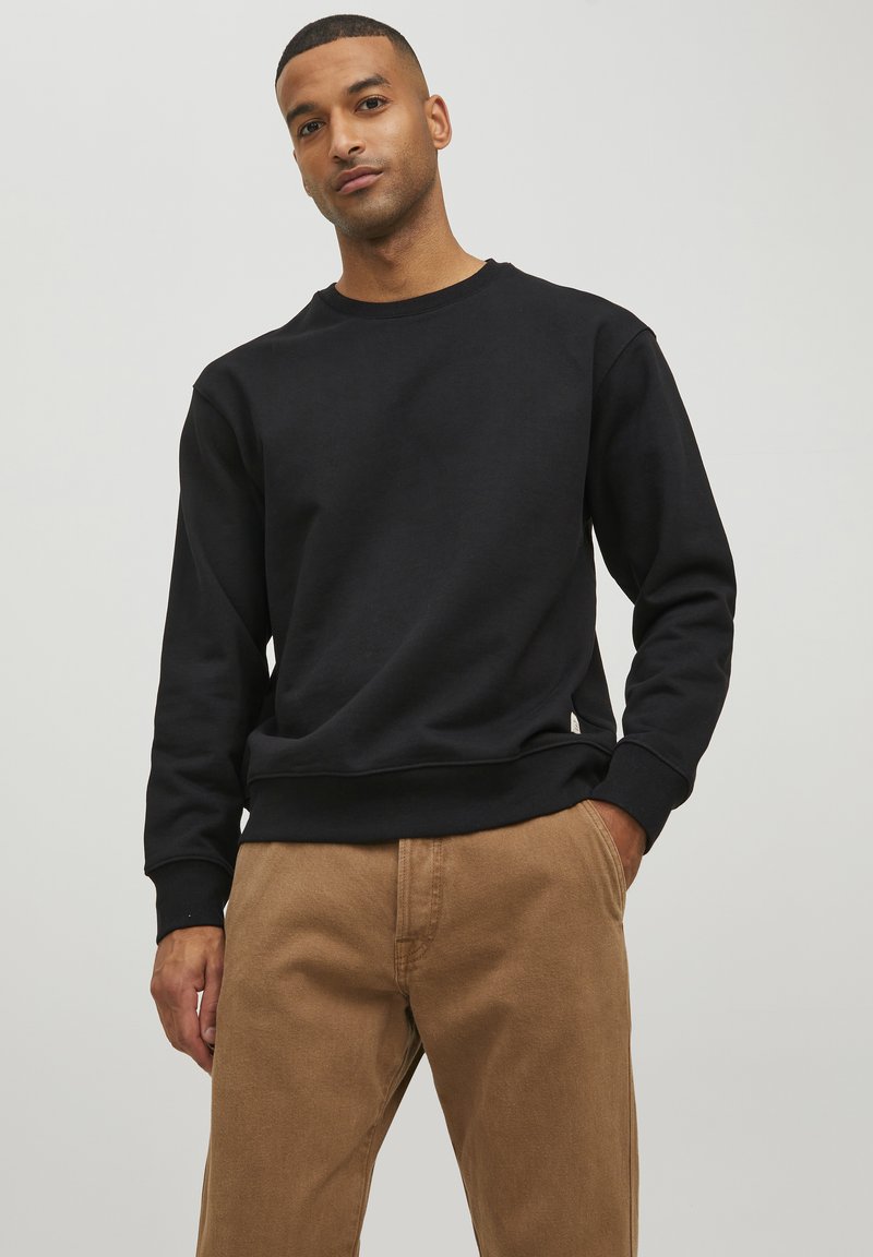 Schwarzer Sweatshirt mit Rundhalsausschnitt, gerippten Bündchen und Saum. Kombiniert mit hellbraunen Hosen. Glatte Textur, lässige Passform, minimales Branding.
