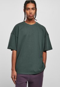 Tricou oversized de culoare verde închis, fabricat din bumbac. Dispune de un guler rotund și mâneci scurte, cu o textură netedă și cusături minime.
