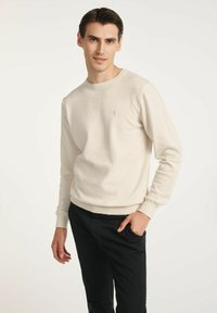 Beżowy sweter dzianinowy z długimi rękawami i prążkowanym wykończeniem, z małym logo na klatce piersiowej, zestawiony z czarnymi spodniami.