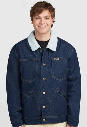 Junger Mann lächelt, trägt eine dunkelblaue Jeansjacke mit weißem Fleecekraagen und O'Neill-Logo auf der Brusttasche.