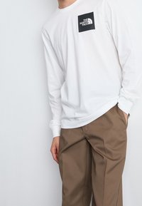 Bílé tričko s dlouhým rukávem a malým černým logem "The North Face" na hrudi, doplněné hnědými kalhotami. Měkká textura, standardní střih, design s kulatým výstřihem.