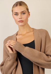 Cardigan marrone a coste con maniche ampie, apertura frontale e tessuto testurizzato. Indossato sopra una maglietta nera, che mostra uno stile di sovrapposizione casual.