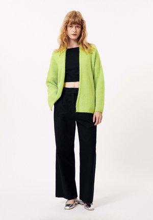 Cardigan texturé vert citron avec une ouverture sur le devant, associé à un top noir court et un pantalon large noir. Chaussures à talons block argentés.