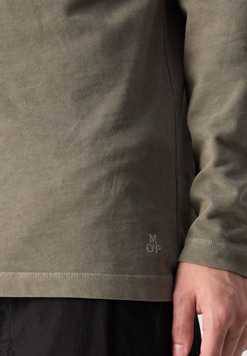Chemise à manches longues vert olive avec une texture lisse, présentant un logo subtil près de l'ourlet et un design à coupe droite.