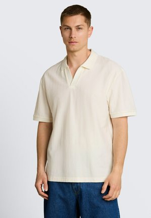 Junger Mann, der ein kurzärmeliges elfenbeinfarbenes, strukturiertes Poloshirt und blaue Jeans trägt, steht vor einem schlichten hellen Hintergrund.