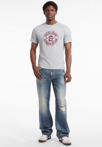 Grå bomulds T-shirt med et cirkulært rødt grafisk print, parret med lyseblå slidte jeans og hvide sneakers med sorte striber.