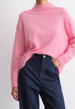 Personne portant un pull en tricot rose pâle avec des poignets et un col côtelés, légèrement rentré dans un jean taille haute bleu foncé.